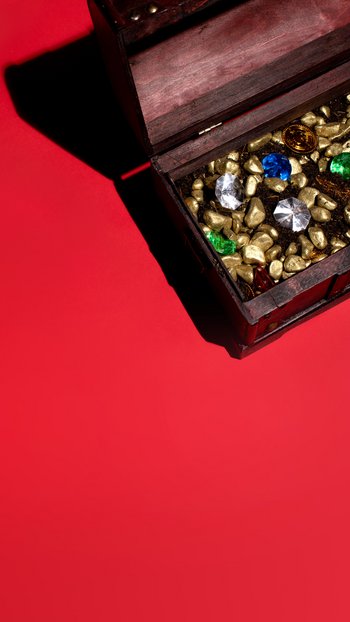 Eine hölzerne Truhe mit einem kleinen innenliegenden Schatz, bestehend aus Diamanten und Goldstückchen, auf einem roten Hintergrund, von oben fotografiert.