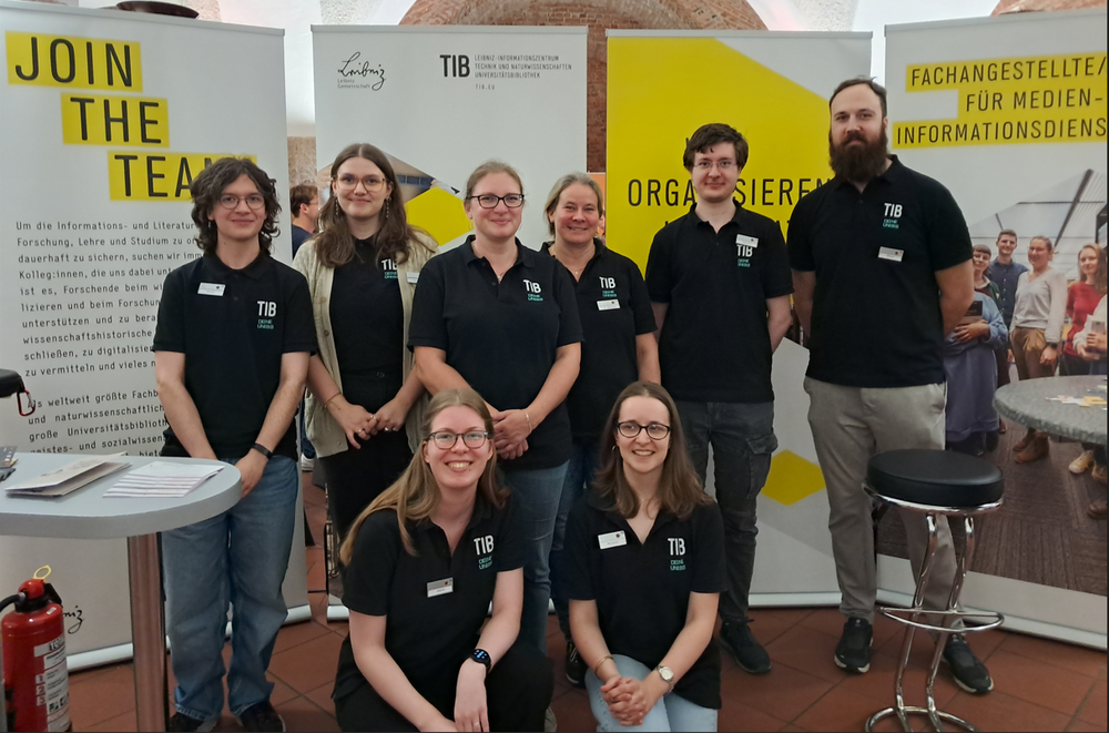 Ein Gruppenbild des TIB-Teams bei der Messe, im Hintergrund sind Aufsteller und Roll-ups des Messestandes zu sehen, alle gucken zu der Kamera.